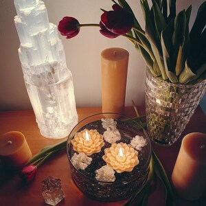 May include: A still life featuring a selenite crystal tower, three candles, a vase of red tulips, and a glass bowl filled with water and floating candles. The candles are all lit and the scene is illuminated by the light from the candles and the selenite crystal.