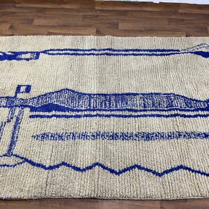 May include: A white rug with a blue geometric pattern. The pattern features a series of horizontal lines and a larger, more intricate design in the centre.