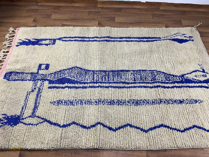May include: A white rug with a blue geometric pattern. The pattern features a series of horizontal lines and a larger, more intricate design in the centre.