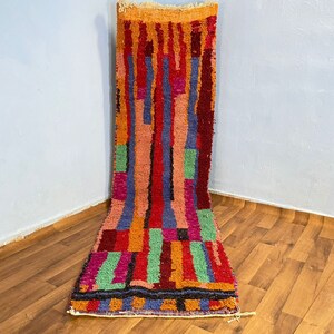 May include: A colorful Moroccan rug with geometric patterns in shades of red, orange, green, blue, and pink. The rug is made of wool and has a hand-woven texture.