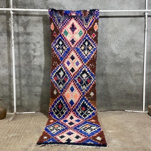 May include: A colorful, patterned rug with a geometric design. The rug features a repeating diamond pattern in shades of brown, blue, pink, and white. The rug is displayed on a white metal bar against a gray concrete wall.