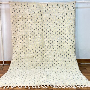 Könnte beinhalten: Cremefarbener Teppich mit einem sich wiederholenden Muster aus kleinen, blauen Strichen. Der Teppich hat eine strukturierte Oberfläche und einen Fransenbesatz am unteren Rand. Der Teppich wird auf einem Holzboden präsentiert.