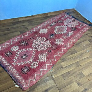 Puede incluir: Una alfombra marroquí vintage de color rosa y blanco con intrincados patrones geométricos. La alfombra tiene un borde con flecos y está hecha de lana.