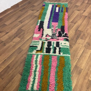 May include: A long, rectangular rug with a colourful geometric pattern. The rug is made of wool and has a soft, plush texture. The colours in the rug include green, pink, white, black, and purple.