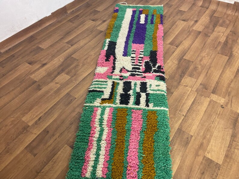 May include: A long, rectangular rug with a colourful geometric pattern. The rug is made of wool and has a soft, plush texture. The colours in the rug include green, pink, white, black, and purple.