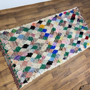 Puede incluir: Una alfombra marroqu&iacute; blanca con un patr&oacute;n de diamantes en tonos de verde, azul, rosa, rojo y negro. La alfombra tiene un borde de flecos.