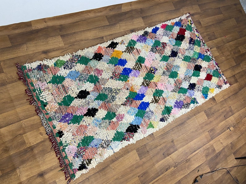 Puede incluir: Una alfombra marroqu&iacute; blanca con un patr&oacute;n de diamantes en tonos de verde, azul, rosa, rojo y negro. La alfombra tiene un borde de flecos.