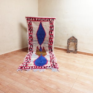 May include: A white, blue, and red Moroccan rug with a geometric pattern. The rug is laid out on a wooden floor.