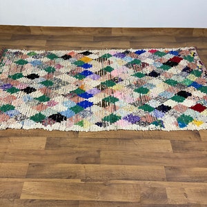 Puede incluir: Una alfombra marroqu&iacute; colorida con un patr&oacute;n de diamantes en tonos de verde, azul, rosa y rojo. La alfombra est&aacute; tejida con un fondo blanco y tiene un aspecto desgastado y vintage.