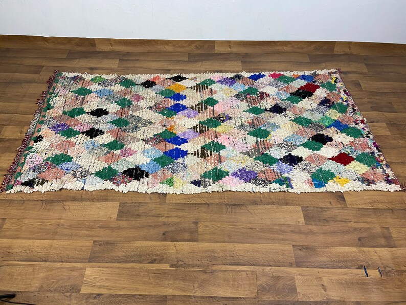 Puede incluir: Una alfombra marroqu&iacute; colorida con un patr&oacute;n de diamantes en tonos de verde, azul, rosa y rojo. La alfombra est&aacute; tejida con un fondo blanco y tiene un aspecto desgastado y vintage.