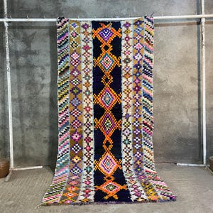 May include: A colorful, patterned rug with a central dark blue stripe featuring diamond shapes in orange and pink. The rug has a variety of colors including pink, yellow, and purple. It is hanging on a white metal rack.