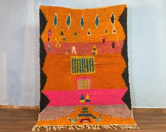 May include: A colorful Moroccan rug with geometric patterns in shades of orange, pink, black, and teal. The rug has a fringe border and is woven with a thick, textured yarn.