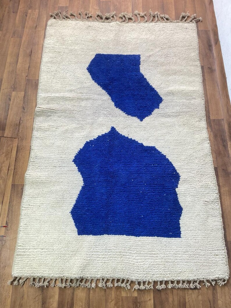 May include: A white rug with two blue abstract shapes. The rug has a fringe border.