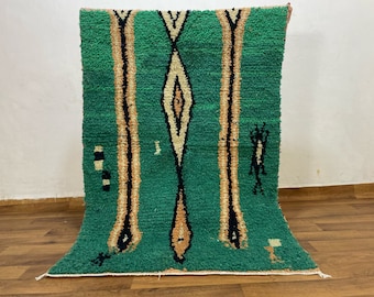 May include: A green and beige Moroccan rug with a geometric design. The rug features a central diamond shape and vertical stripes in beige, black, and green. The rug is made of a thick, textured material.