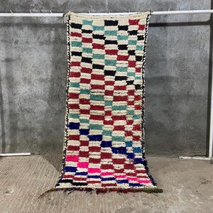 Puede incluir: Una alfombra rectangular tejida a mano con un patrón de cuadros en rojo, negro, azul, turquesa, rosa y blanco. La alfombra se exhibe contra una pared de hormigón gris y está sostenida por un soporte de metal blanco.