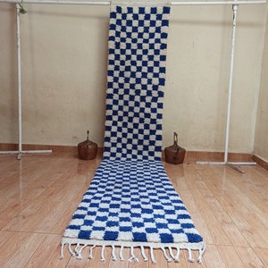 Può includere: Un tappeto lungo a scacchi bianchi e blu con un bordo a frangia. Il tappeto è appeso a un rack di metallo bianco.