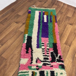 May include: A long, narrow rug with a colourful geometric pattern. The rug is made of wool and has a fringe on both ends. The colours in the rug include pink, green, purple, black, and white.