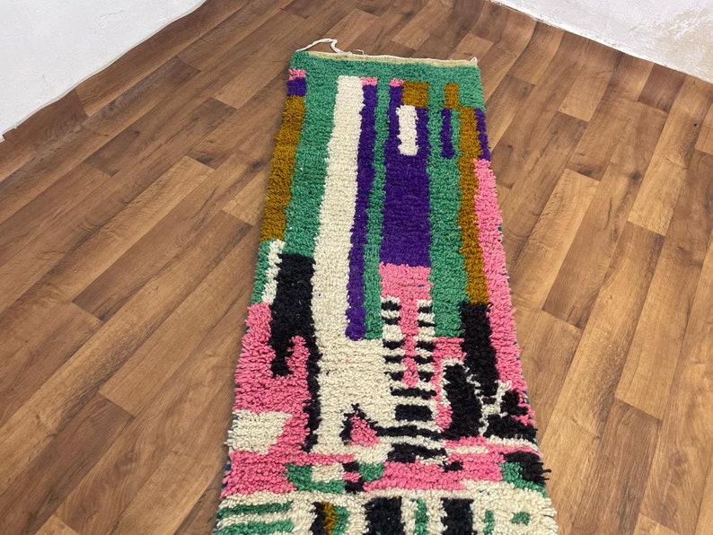 May include: A long, narrow rug with a colourful geometric pattern. The rug is made of wool and has a fringe on both ends. The colours in the rug include pink, green, purple, black, and white.