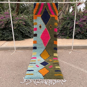 May include: A colorful, geometric rug with a vibrant design. The rug features a combination of blue, orange, pink, and green hues, with diamond and stripe patterns. It is displayed outdoors, showcasing its texture and design.