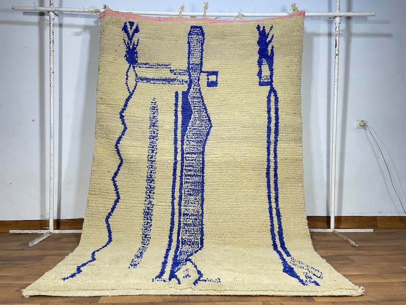 May include: A beige rug with a blue abstract design. The design features two tall, thin shapes with a smaller shape at the top. The rug has a fringe border.