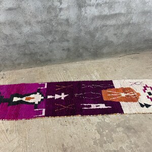 Peut inclure: Un long tapis rectangulaire au design vibrant. Le tapis présente des sections de violet profond, de rose vif, d'orange, de noir et de blanc, avec des motifs géométriques. Le tapis est posé sur un sol en béton.