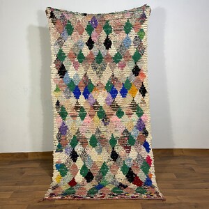 Puede incluir: Una alfombra marroqu&iacute; colorida con un patr&oacute;n de diamantes en tonos de rosa, verde, azul y negro. La alfombra est&aacute; tejida con un hilo grueso y texturizado y tiene flecos en los extremos.