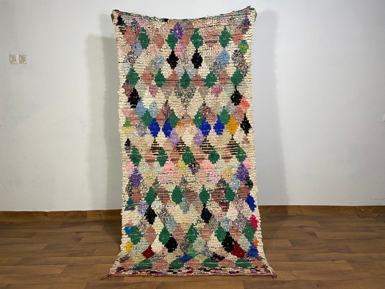 Puede incluir: Una alfombra marroqu&iacute; colorida con un patr&oacute;n de diamantes en tonos de rosa, verde, azul y negro. La alfombra est&aacute; tejida con un hilo grueso y texturizado y tiene flecos en los extremos.