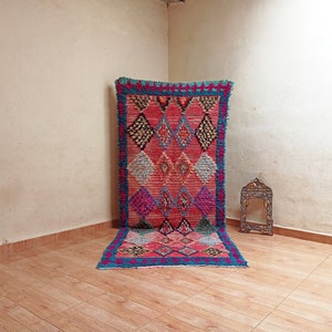 Peut inclure: Un tapis rectangulaire vibrant avec un motif de losanges dans les tons de rose, de bleu et de noir. Le tapis a un aspect texturé et une bordure à franges. Le tapis est présenté contre un mur neutre.
