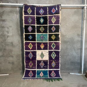 Peut inclure: Un tapis rectangulaire tissé à la main avec un motif géométrique. Le tapis présente une grille de carrés dans les tons de violet, noir et sarcelle, chacun contenant un motif de losange. Le tapis a une bordure blanche et une frange verte en bas. Le tapis est suspendu à une barre métallique blanche.