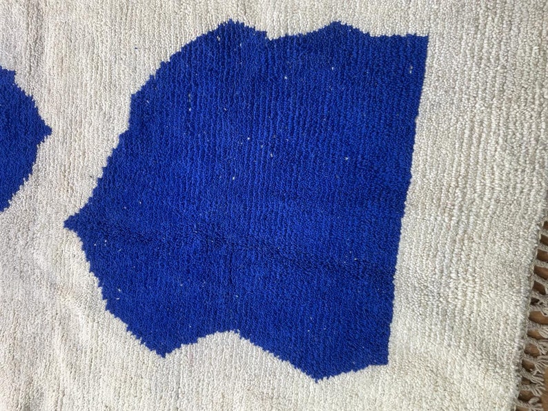 May include: A white rug with a blue abstract design. The rug has a fringe border.