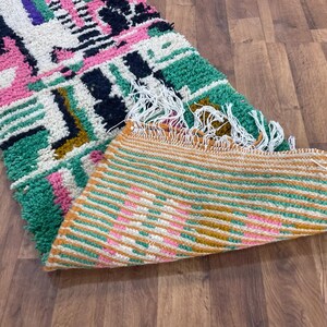 May include: A colourful Moroccan rug with geometric patterns in shades of pink, green, black, and white. The rug has a fringe border and is folded in half, revealing a striped pattern on the underside.