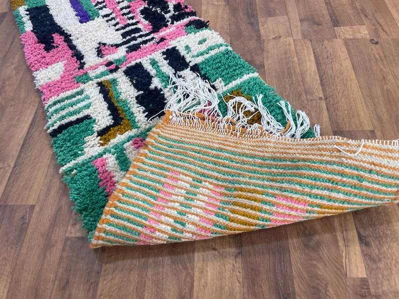May include: A colourful Moroccan rug with geometric patterns in shades of pink, green, black, and white. The rug has a fringe border and is folded in half, revealing a striped pattern on the underside.