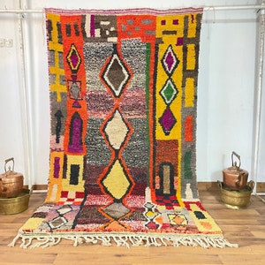 May include: A colorful Moroccan rug with geometric patterns in shades of red, orange, yellow, green, purple, and brown. The rug has a fringe border and is hanging on a white wall.