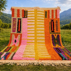 Peut inclure: Un tapis rectangulaire coloré avec un motif géométrique. Le tapis présente une variété de couleurs, notamment du rose, de l'orange, du jaune et du violet. Il a une bordure à franges et est présenté à l'extérieur sur un champ herbeux.