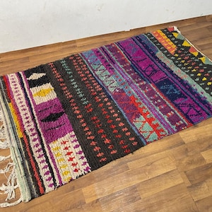 Op de afbeelding: Een kleurrijk, handgeweven tapijt met een geometrisch patroon in tinten rood, paars, blauw, geel en zwart. Het tapijt heeft een franje rand en is gemaakt van wol.