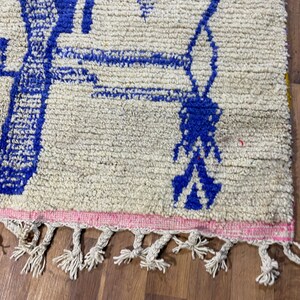 May include: A white rug with a blue geometric design and pink fringe. The rug has a white background with a blue geometric design. The rug has a pink fringe around the edges.