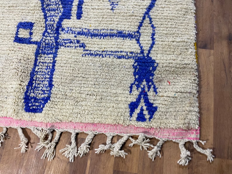 May include: A white rug with a blue geometric design and pink fringe. The rug has a white background with a blue geometric design. The rug has a pink fringe around the edges.