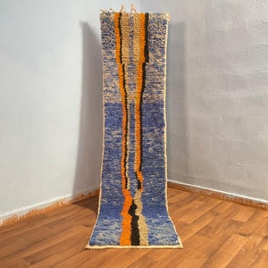 May include: A long, narrow rug with a blue, orange, and black geometric pattern. The rug is woven with a thick, textured yarn and has a fringe on both ends.