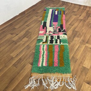 May include: A long, rectangular rug with a geometric pattern in shades of green, pink, brown, white, and black. The rug has a fringe on both ends.