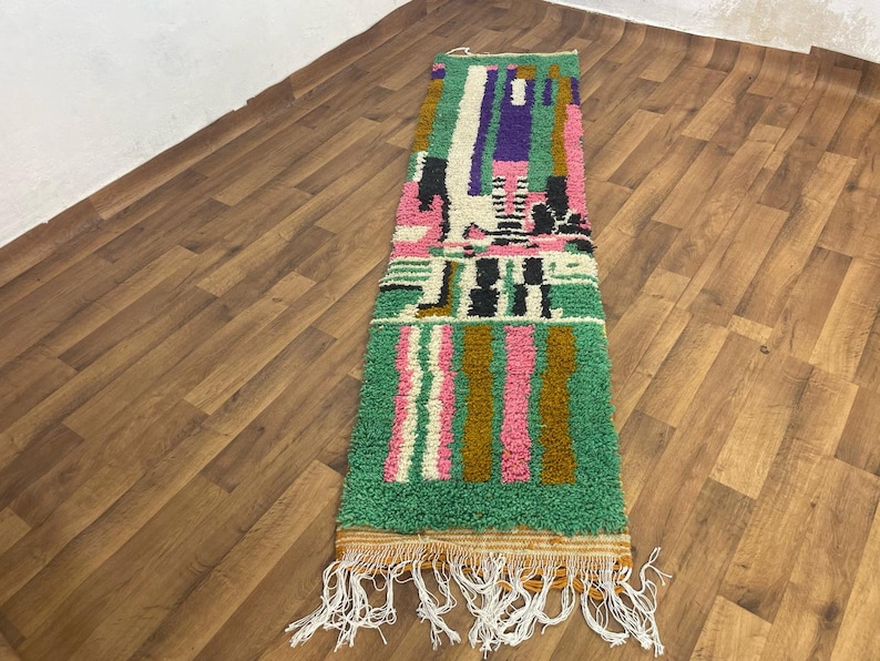 May include: A long, rectangular rug with a geometric pattern in shades of green, pink, brown, white, and black. The rug has a fringe on both ends.