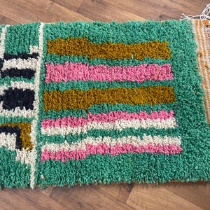 May include: A green, pink, white and gold patterned rug with fringe. The rug has a geometric design.