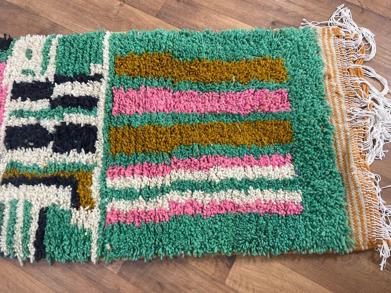 May include: A green, pink, white and gold patterned rug with fringe. The rug has a geometric design.