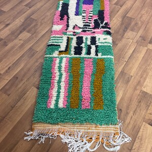 May include: A long, rectangular rug with a geometric pattern in shades of green, pink, white, black, and brown. The rug has a fringe border.