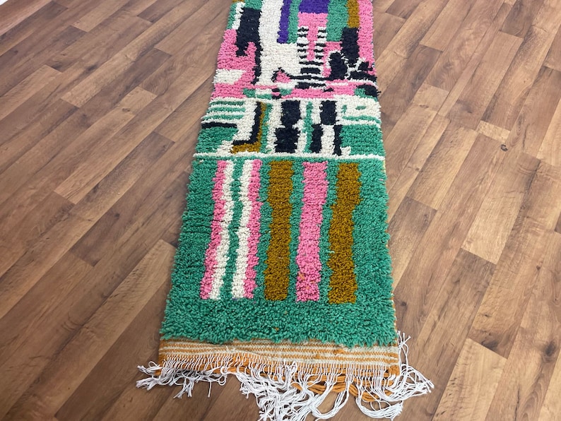May include: A long, rectangular rug with a geometric pattern in shades of green, pink, white, black, and brown. The rug has a fringe border.