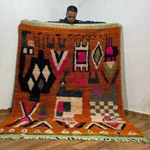 Peut inclure: Un tapis tissé à la main aux couleurs vives, avec un fond orange brûlé et des motifs géométriques noirs, blancs, roses et verts. Le tapis présente des formes de diamants, de triangles et de carrés, avec une frange verte sur le bord inférieur. Tapis de style marocain.