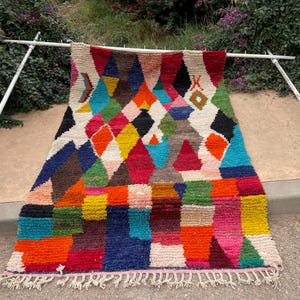 Peut inclure: Un tapis coloré avec des motifs géométriques en losanges et rectangulaires. Le tapis présente une variété de couleurs, dont le rouge, le bleu, le jaune et le vert. Il est exposé à l'extérieur, suspendu à un cadre métallique blanc.