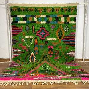 May include: A colourful, hand-woven rug with a bright green background and geometric patterns. The rug features pink, blue, orange, and white accents, with a fringed edge. The rug is displayed against a white wall and wooden floor.