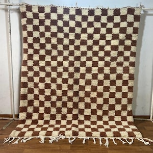 Può includere: Un tappeto a scacchi marroni e bianchi con un bordo a frangia. Il tappeto è realizzato in un materiale morbido e soffice e presenta un design tradizionale marocchino.