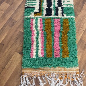 May include: A green, pink, white and brown striped rug with a geometric pattern. The rug has a fringe border.