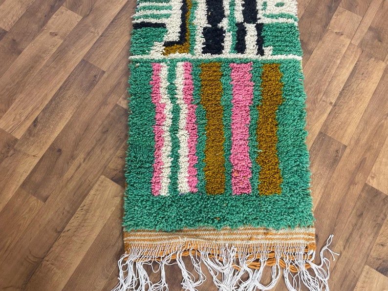 May include: A green, pink, white and brown striped rug with a geometric pattern. The rug has a fringe border.